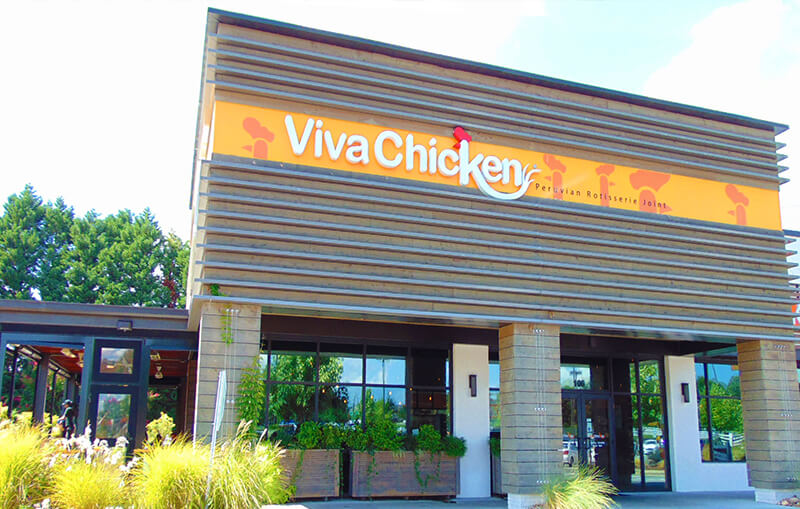 Exterior of a Viva Chicken restaurant with a bright orange sign and modern wooden facade, large windows, greenery in front, and clear blue sky in the background.