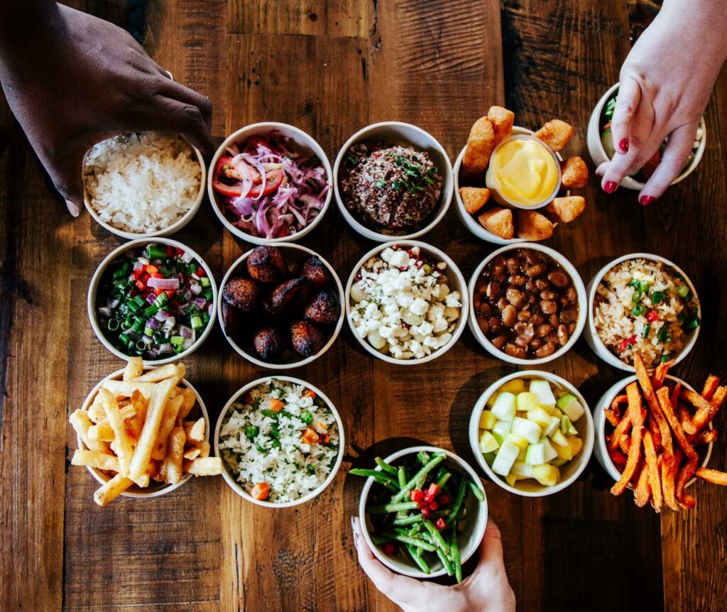 Three hands reach for an assortment of small bowls filled with various Viva Chicken foods, such as rice, beans, fries, salads, fried plantains, sauces, and pickled vegetables, arranged on a wooden table.