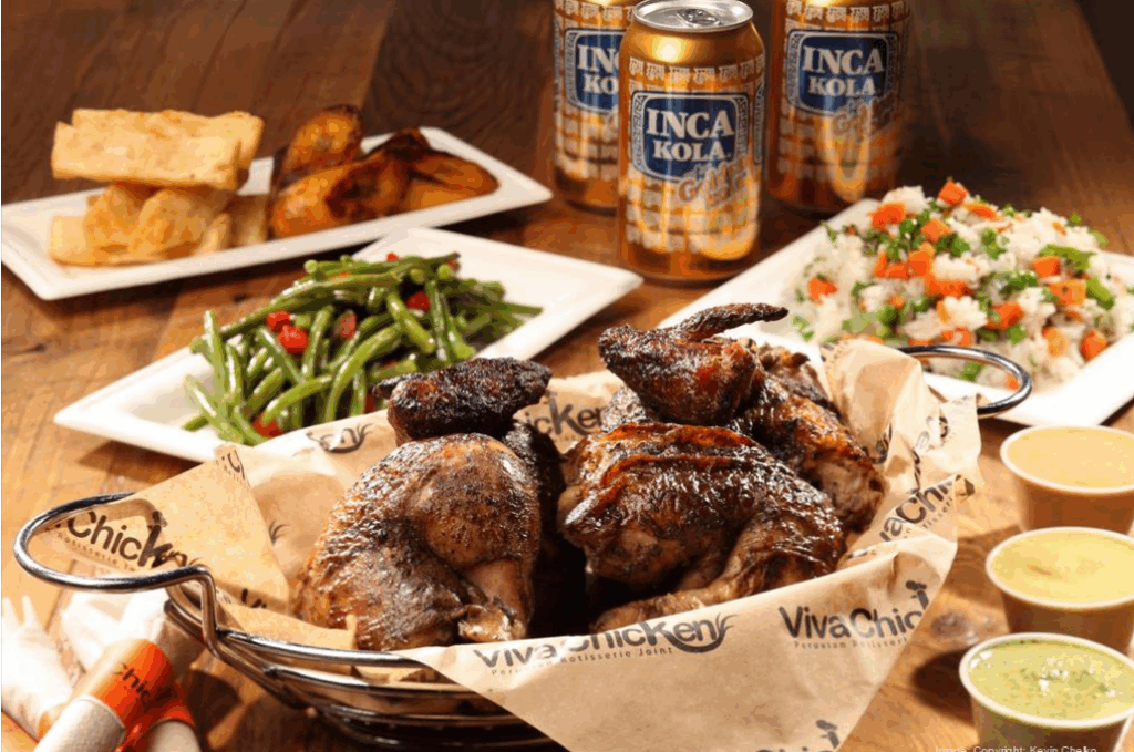 A table with roasted chicken, green beans, potato wedges, white rice with vegetables, sauces, and three cans of Inca Kola, served on Viva Chicken-branded paper.