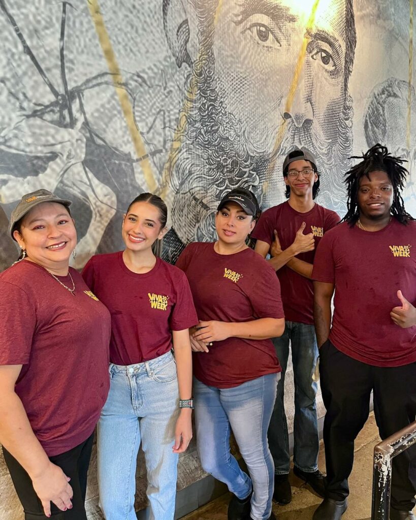 Five people wearing matching maroon shirts and standing in front of a large mural featuring a black-and-white portrait. They are smiling and posing for the photo indoors.