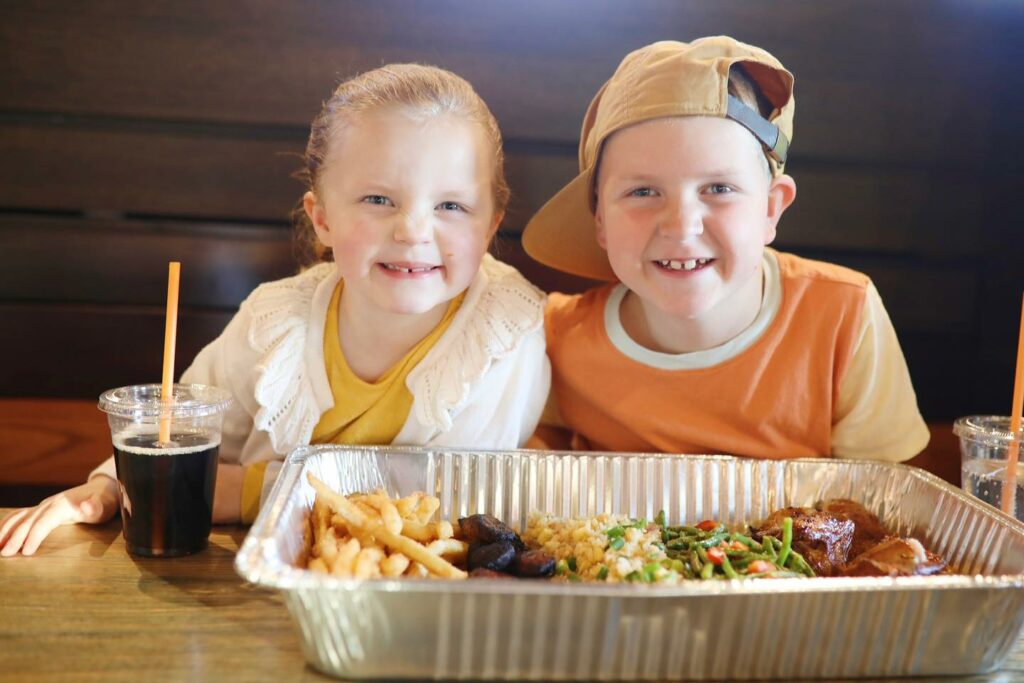 Two smiling children sit at a table with a large tray of food, including fries, greens, and other items. Both have drinks with orange straws; one child wears a backward cap and the other has braided hair.