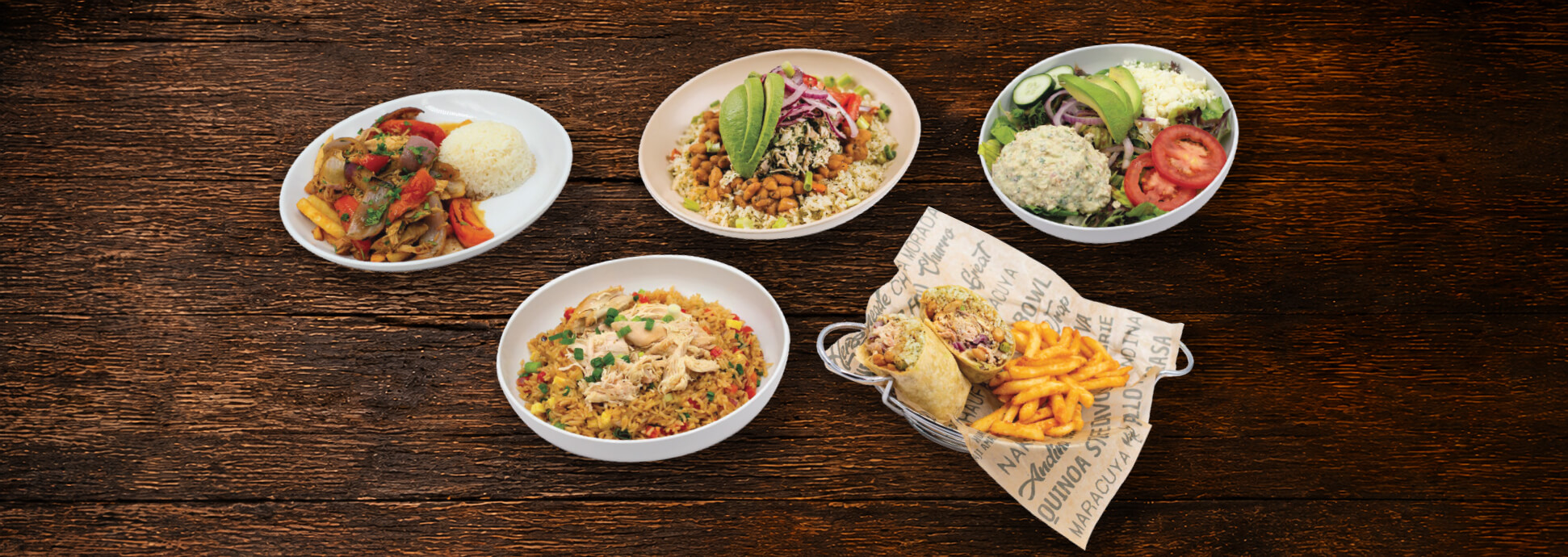Five plates of food on a wooden table, featuring rice dishes with vegetables, a salad with tomato and cucumber, a bowl with avocado slices, and a sandwich wrap served with fries in a basket.